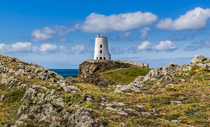 A white lighthouse stands on a rocky outcrop overlooking the sea under a blue sky with white clouds.